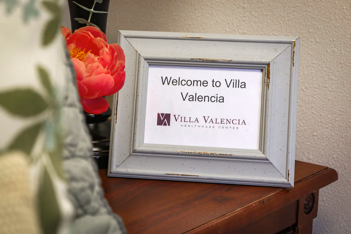 A framed sign on a wooden table that reads 'Welcome to Villa Valencia' with the Villa Valencia Healthcare Center logo below. There is a red flower in a vase partially visible behind the frame and a blurred patterned cushion in the foreground.
