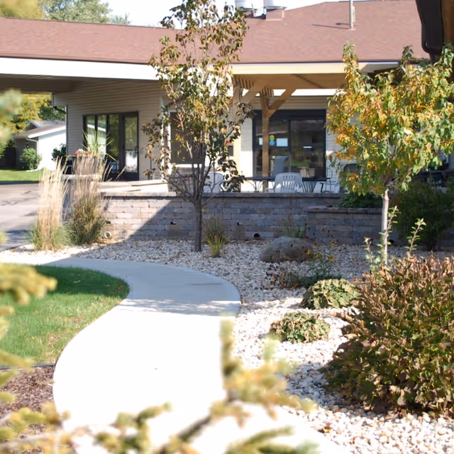 A curved concrete path leads through a rock garden and landscaping to the covered entrance and patio of a single-story senior living building.