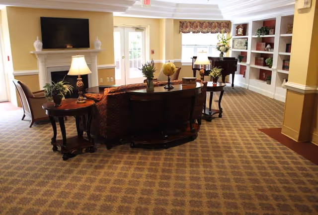 A cozy living room area in a senior living facility with patterned carpet, a sofa, side tables with lamps and plants, a fireplace with a mounted flat-screen TV above it, a grand piano near a window with floral valance, and built-in bookshelves filled with books and decorative items.