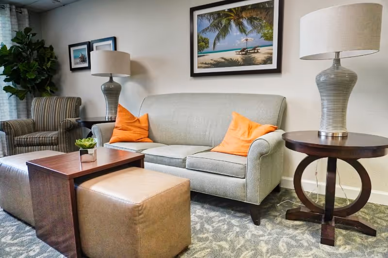 A cozy living room area with a light gray sofa adorned with two orange pillows, a striped armchair, a wooden coffee table with a small plant, a beige ottoman, two matching table lamps on wooden side tables, and framed beach-themed artwork on the wall.