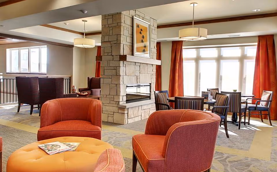 Spacious senior living common area with red upholstered chairs, a central stone fireplace, round tables and large windows.
