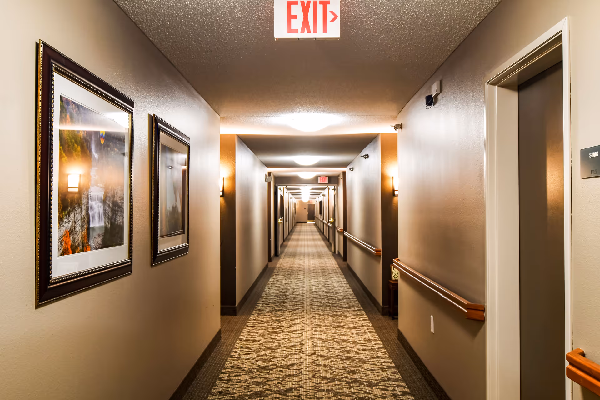A long, well-lit hallway in a senior living facility with framed pictures on the left wall, handrails on both sides, and multiple doors along the corridor. An illuminated exit sign is visible on the ceiling.