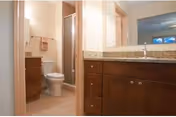 Interior view of a senior living community bathroom featuring a toilet, a glass-enclosed shower, and a wooden vanity with a granite countertop and a sink. The walls are light-colored, and there is a towel hanging near the toilet.