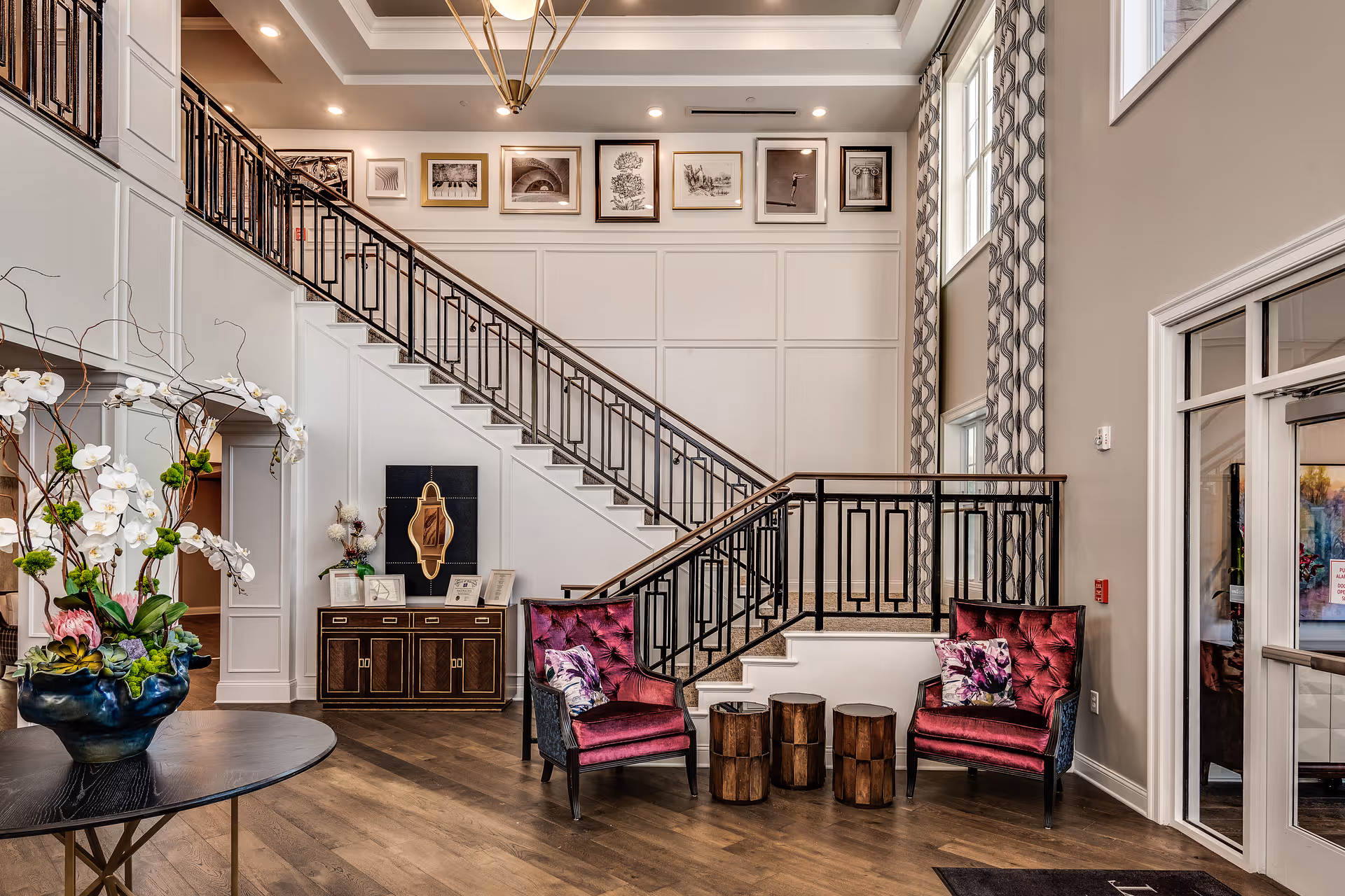 A stylish interior lobby area featuring a staircase with black metal railings, two plush magenta armchairs with floral pillows, three small wooden stump side tables, a dark wood console table with decorative items, and a round table with a large floral arrangement. The walls are decorated with framed artwork and large windows with patterned curtains allow natural light to fill the space.