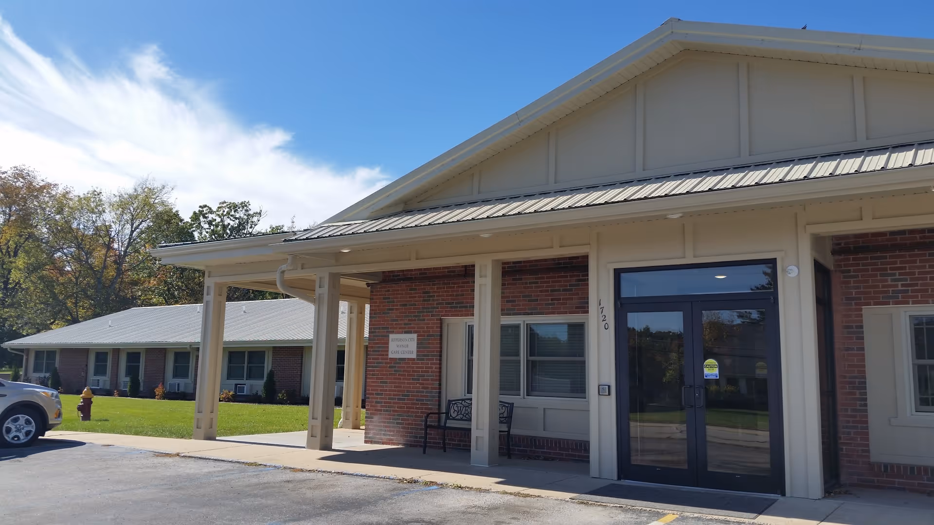 Exterior view of Jefferson City Manor showing the entrance with a covered porch supported by columns, a bench near the door, and a brick and beige siding facade. A car is parked on the left side and there is a grassy area with trees in the background under a partly cloudy sky.