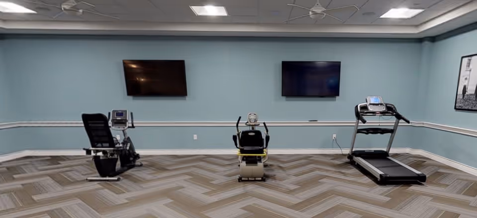 A small exercise room with light blue walls and patterned carpet flooring. The room contains three pieces of exercise equipment: a recumbent bike on the left, a stationary bike in the center, and a treadmill on the right. Two flat-screen TVs are mounted on the wall above the exercise machines. A framed black and white picture is hung on the right wall.