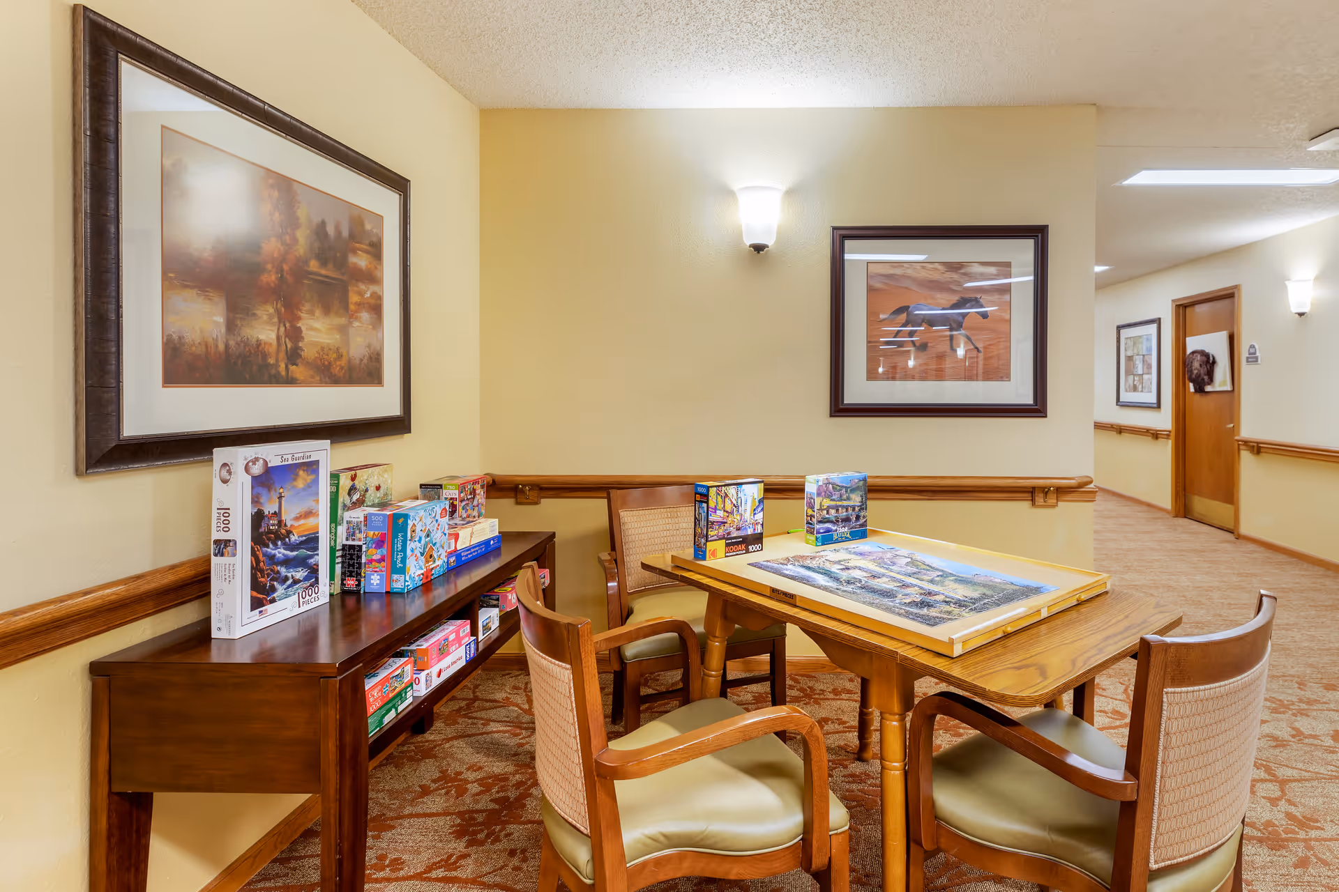 A cozy activity corner in a senior living facility with a wooden table and four chairs. On the table, there is a partially completed jigsaw puzzle and several puzzle boxes. A wooden console table against the wall holds more puzzle boxes. The walls are decorated with framed artwork, and the area is softly lit by wall sconces. The carpet has a floral pattern, and a hallway with additional artwork is visible in the background.