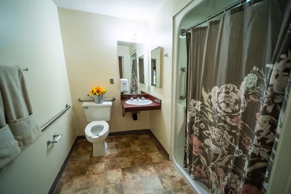 A bathroom with a toilet, a sink with a maroon countertop, a mirror above the sink, and a shower with a floral-patterned curtain. There are grab bars near the toilet and shower, a towel hanging on the wall, and a small vase with yellow flowers on the toilet tank.