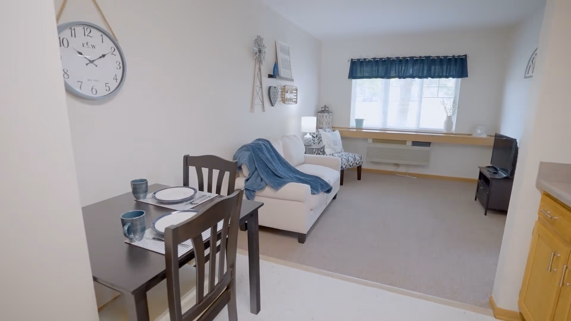 A cozy living room and dining area in a senior living facility. The dining area has a small dark wooden table set for two with blue mugs and white plates. The living room features a white couch with a blue throw blanket, a patterned armchair, a small TV on a stand, and a large window with blue curtains letting in natural light. The walls are decorated with a large clock and various wall hangings.