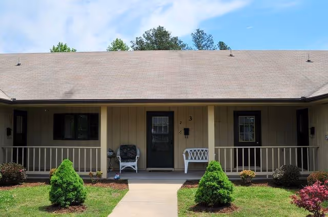 Front porch of a single-story senior living building with a central door, two chairs on the covered porch, and small shrubs lining a concrete walkway.