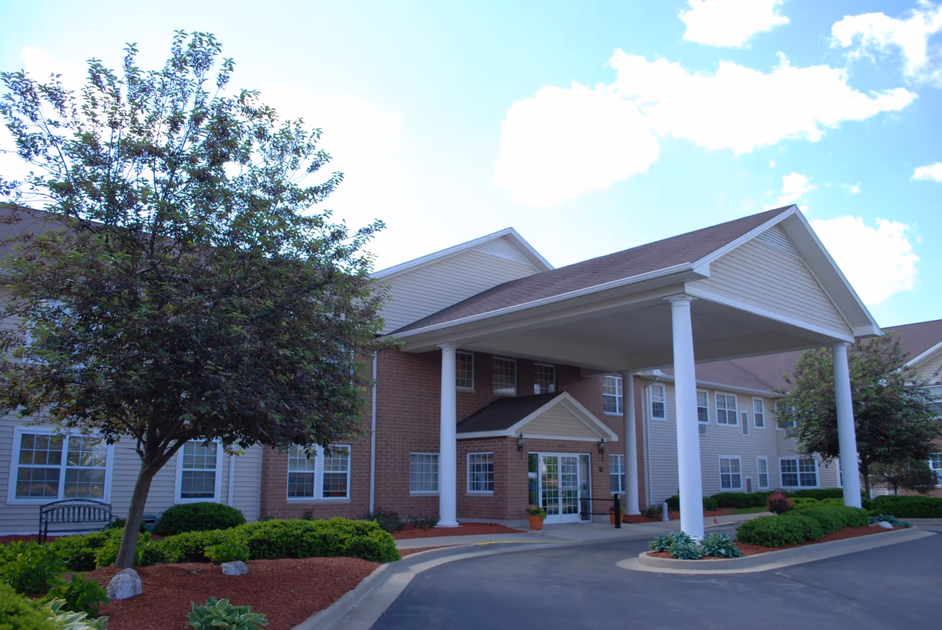 Exterior view of Bowman Estates of Danville, showing the front entrance with a covered driveway supported by white columns, surrounded by landscaped bushes, trees, and a clear blue sky with some clouds.