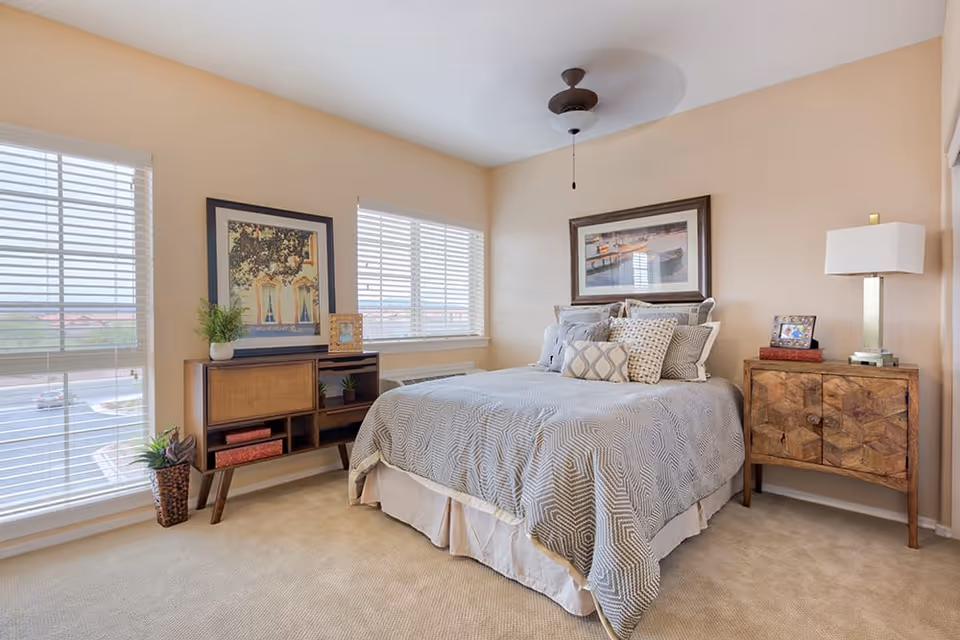 A bright and cozy bedroom with a large bed covered in a gray patterned comforter and multiple pillows. The room features beige walls, two large windows with white blinds, a wooden nightstand with a lamp and picture frame, and a wooden console table with plants and decorative items. A ceiling fan is mounted above the bed.