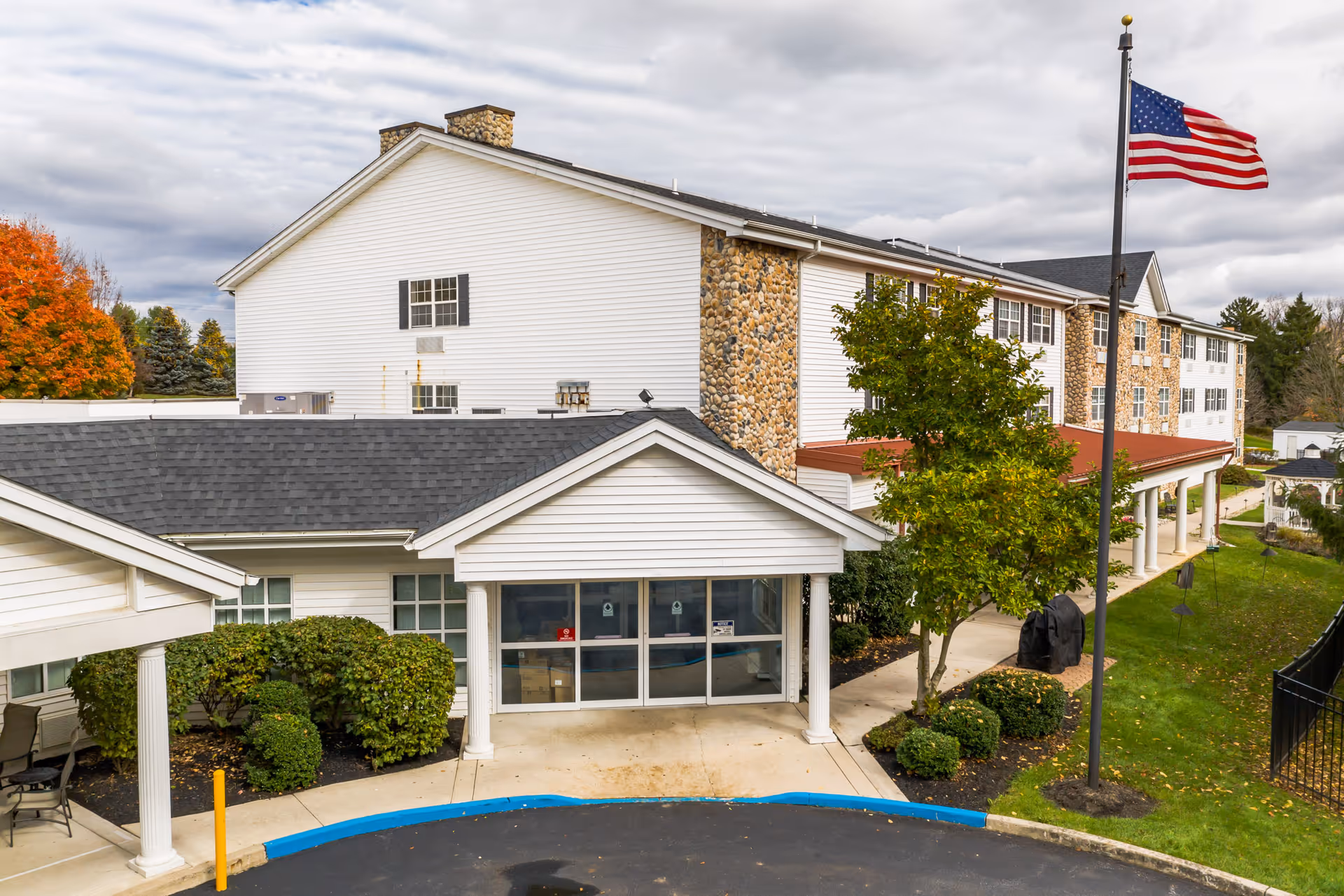 Front exterior of a multi-story senior living facility with white siding and stone accents, a covered entrance, landscaping, and an American flag.