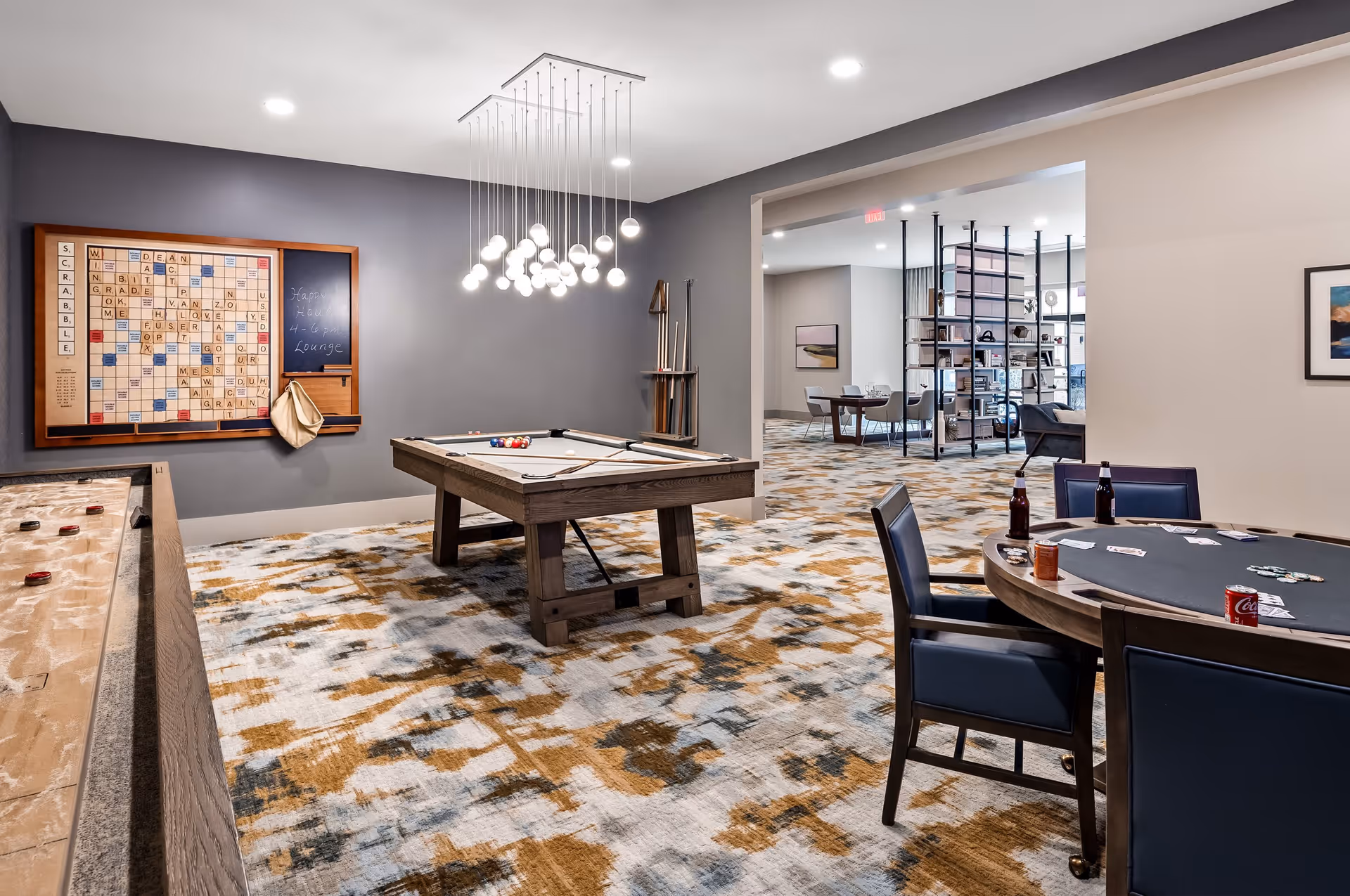 A recreational lounge area featuring a pool table with billiard balls and cues, a large Scrabble board mounted on the wall, a shuffleboard table, and a round card table with chairs. The room has modern lighting fixtures and a patterned carpet. In the background, there is an open space with additional seating and shelving.