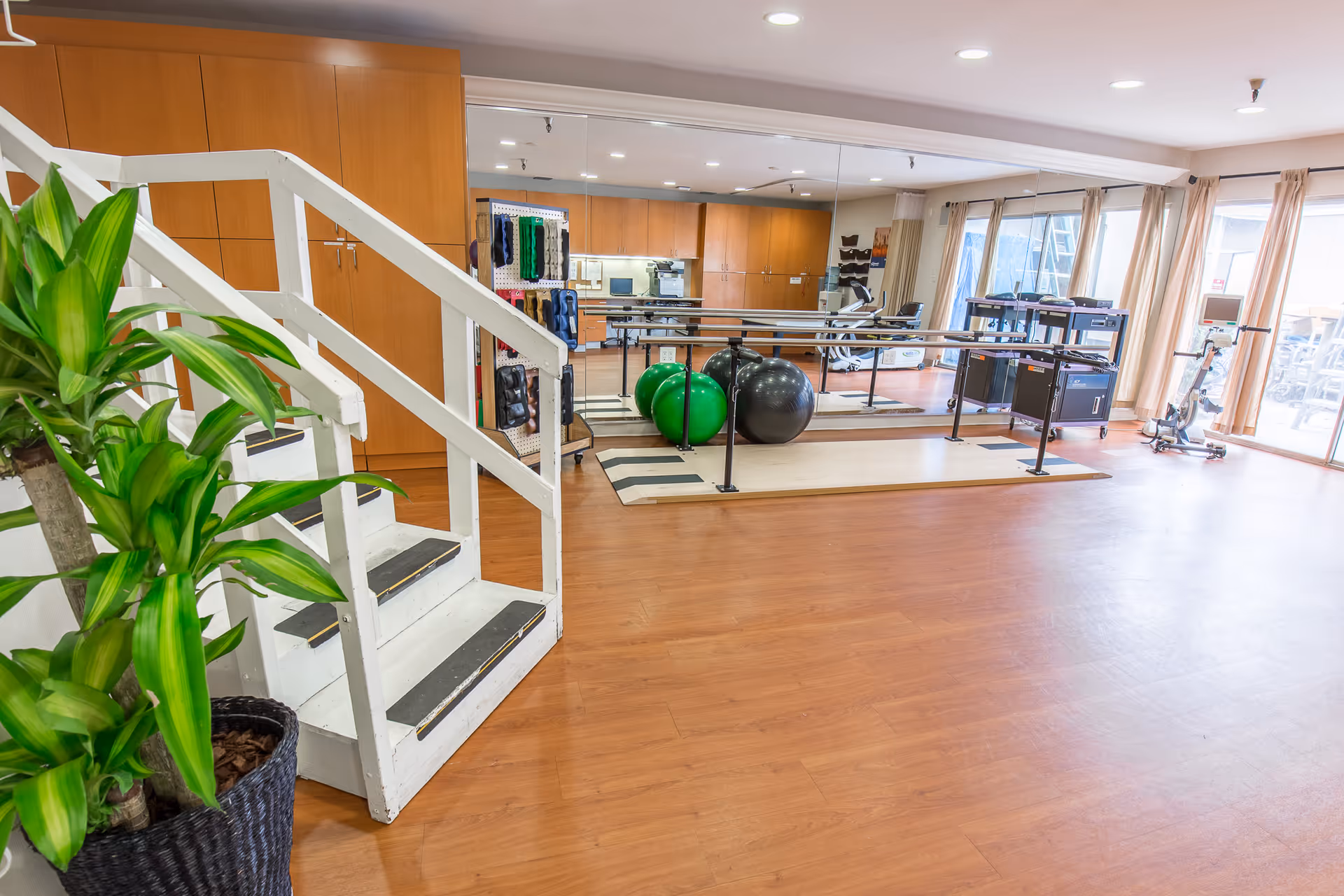 A spacious rehabilitation or exercise room with wooden flooring, a white wooden staircase, large mirrors on the wall, exercise balls, parallel bars for walking practice, and various exercise equipment. There are large windows with beige curtains allowing natural light to enter the room.