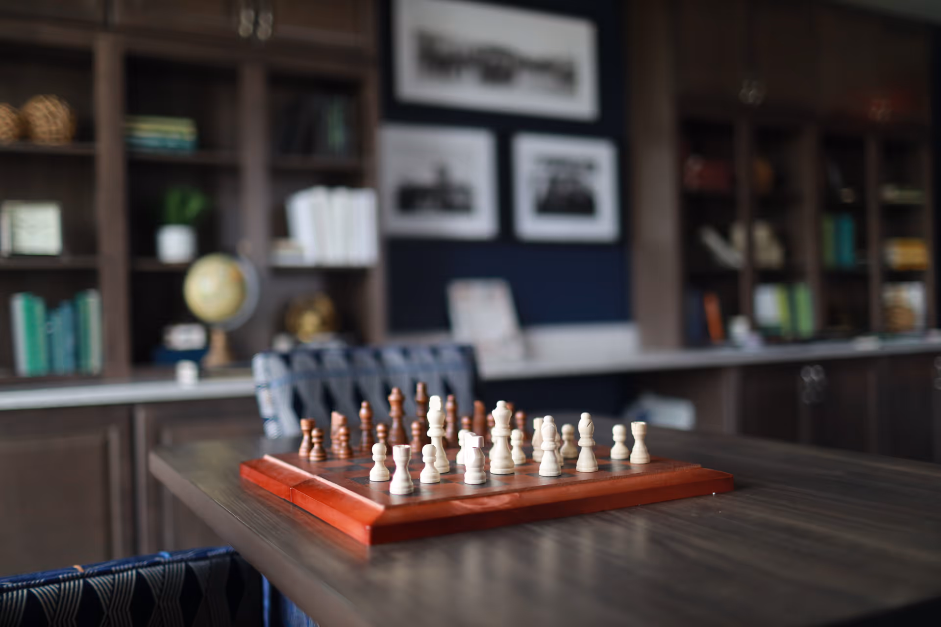 A chessboard with chess pieces set up on a wooden table in a room with dark wood bookshelves filled with books and decorative items in the background.