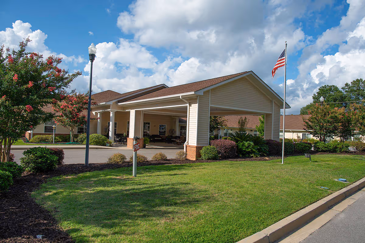 Exterior view of Spring Oak at Lexington senior living facility showing the entrance with a covered driveway, landscaped lawn with bushes and trees, an American flag on a flagpole, and a partly cloudy sky.