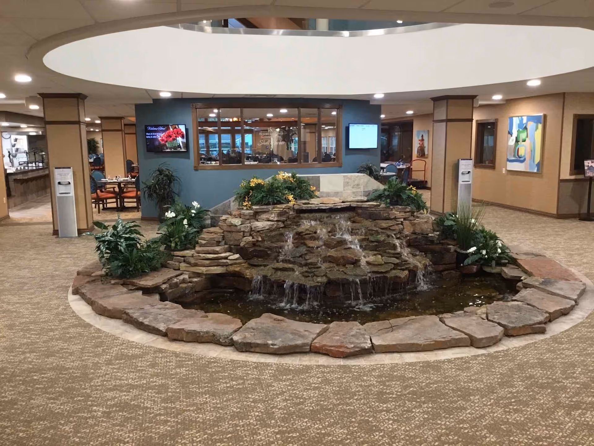 Indoor area of Shell Harbor Retirement Community featuring a central stone water fountain with cascading water surrounded by plants. The space has carpeted floors, beige walls with artwork, and seating areas visible through a large window in the background.