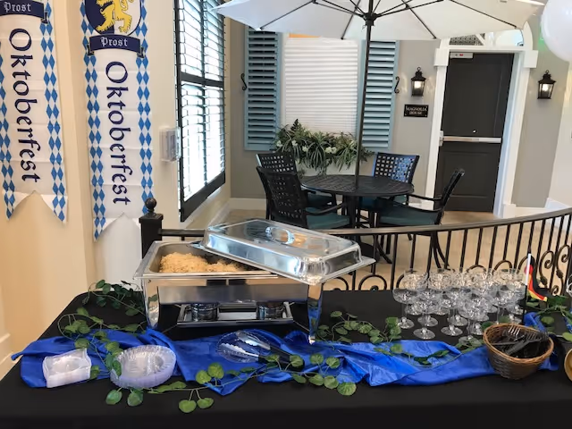 Indoor event setup with a buffet chafing dish containing food, a table covered with a black cloth and blue runner decorated with green leaves, empty wine glasses, and utensils in a basket. Two Oktoberfest banners hang on the wall to the left. In the background, there is a round table with four chairs and a large white umbrella, next to a door labeled Magnolia Trails at East Lake.