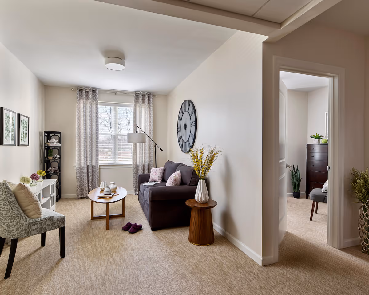 A cozy living room with beige walls and carpet, featuring a dark gray sofa with pink pillows, a wooden coffee table with decorative items, a patterned armchair with a cushion, a tall black shelving unit, and a large wall clock. The room has a window with patterned curtains letting in natural light. An open doorway leads to a bedroom with a dark wooden dresser and a gray chair.