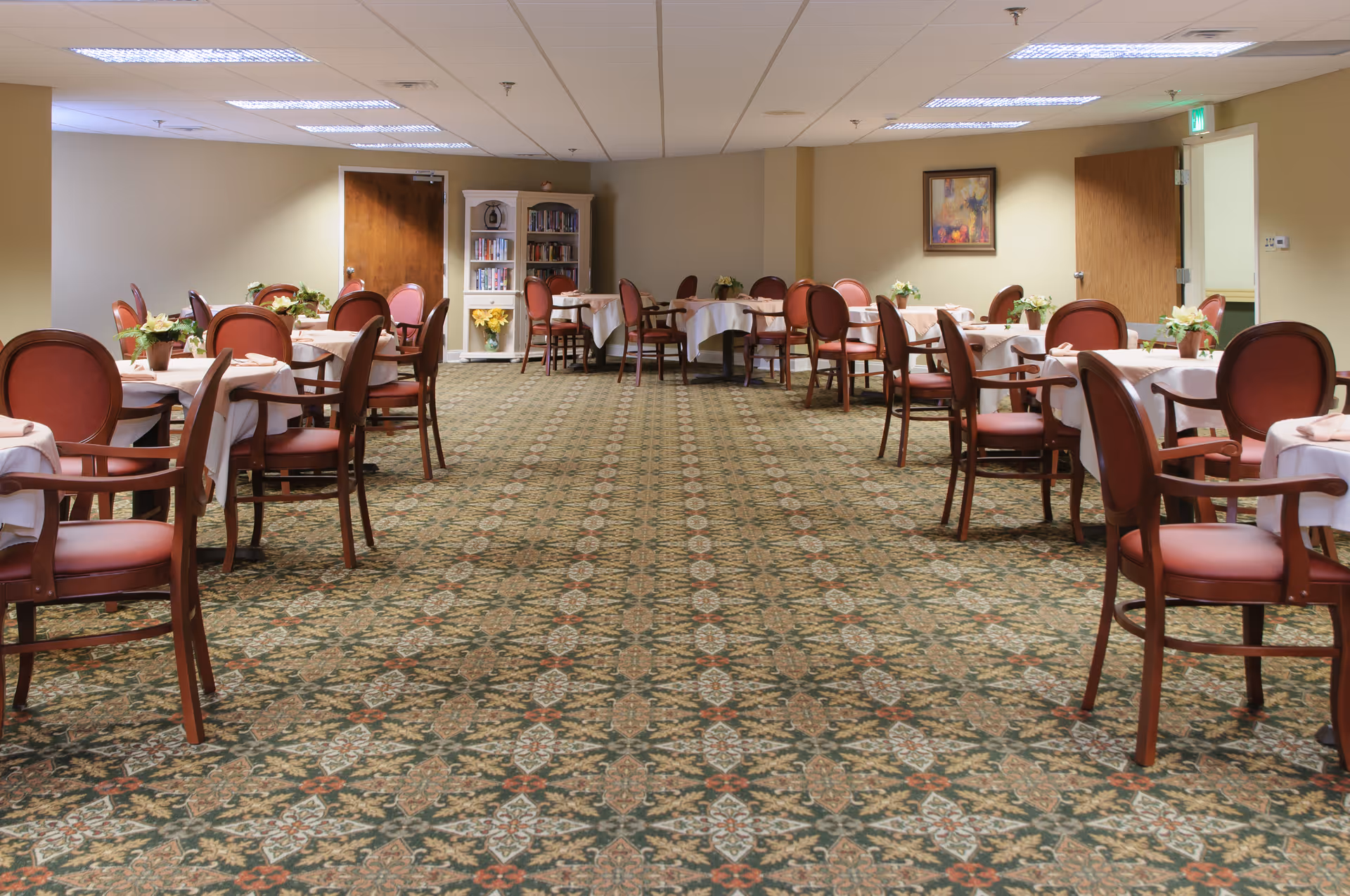 A spacious dining room with multiple round tables covered with white tablecloths and surrounded by wooden chairs with red cushions. The room has patterned carpet flooring, beige walls, fluorescent ceiling lights, and a bookshelf with books and decorative items against the far wall. There are two wooden doors and a framed picture on the wall.