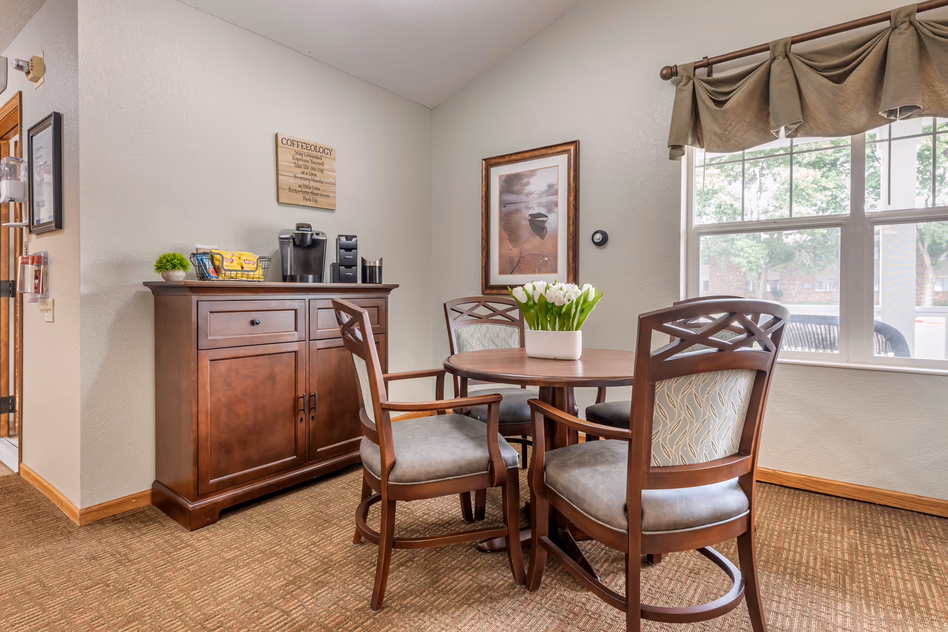 A cozy seating area with a round wooden table and four cushioned chairs with wooden frames. On the table is a white vase filled with white tulips. Behind the table is a wooden cabinet with a coffee maker, coffee supplies, and a small green plant on top. A framed picture of a boat on water hangs on the wall above the cabinet, and a window with a valance lets in natural light, showing trees outside.