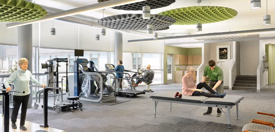 A spacious fitness and rehabilitation room in a senior living facility with exercise equipment including treadmills and a stationary bike. An elderly woman is using parallel bars for walking support, two seniors are exercising on machines near large windows, and a staff member is assisting an elderly woman with leg stretches on a therapy table. The room has modern ceiling fixtures and a carpeted floor.