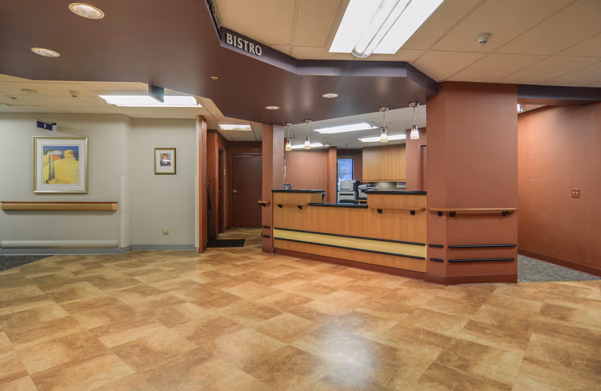 Interior view of a reception or check-in area labeled 'Bistro' with a wooden counter, overhead lights, and a tiled floor. The walls are painted in warm tones with handrails and framed artwork visible on the left side.