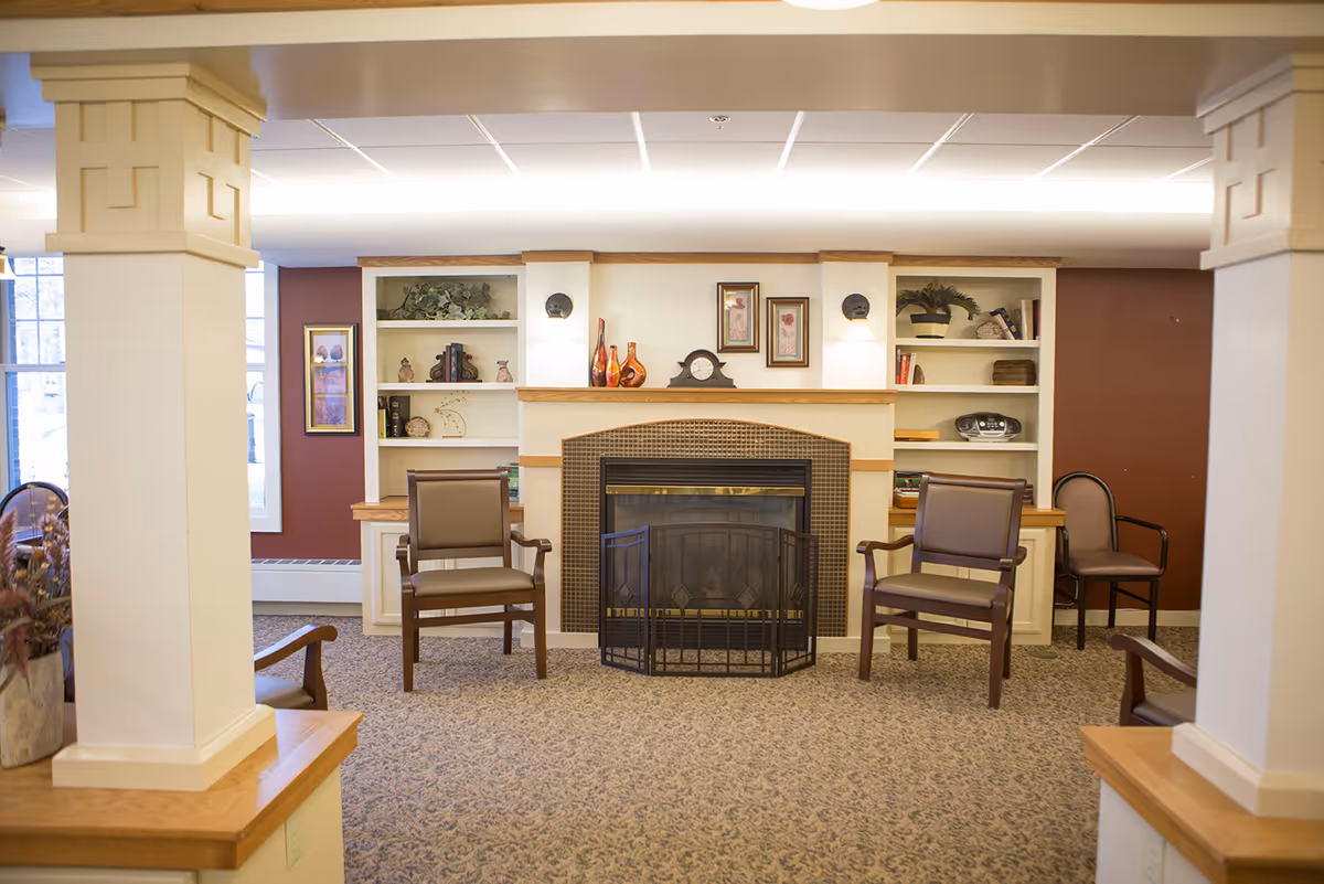 A cozy common area with a fireplace in the center, flanked by built-in shelves decorated with vases, books, and plants. Two armchairs are placed in front of the fireplace, with additional chairs along the walls. The room has carpeted flooring and is softly lit with ceiling lights.