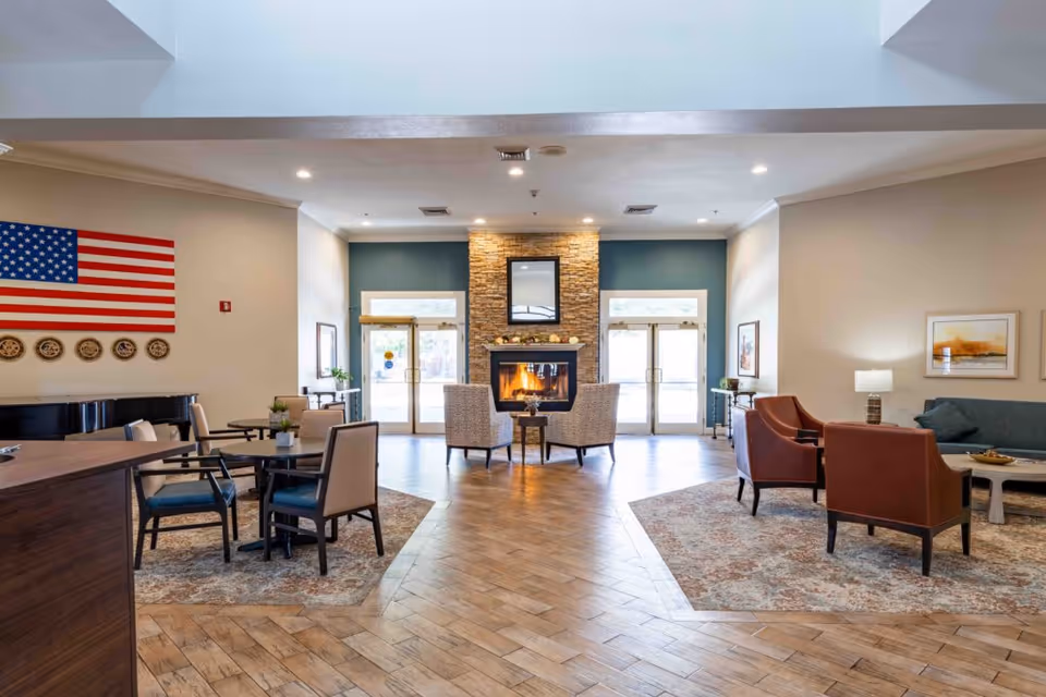Spacious community lounge with seating areas, a central stone fireplace, tables, and an American flag on the wall.