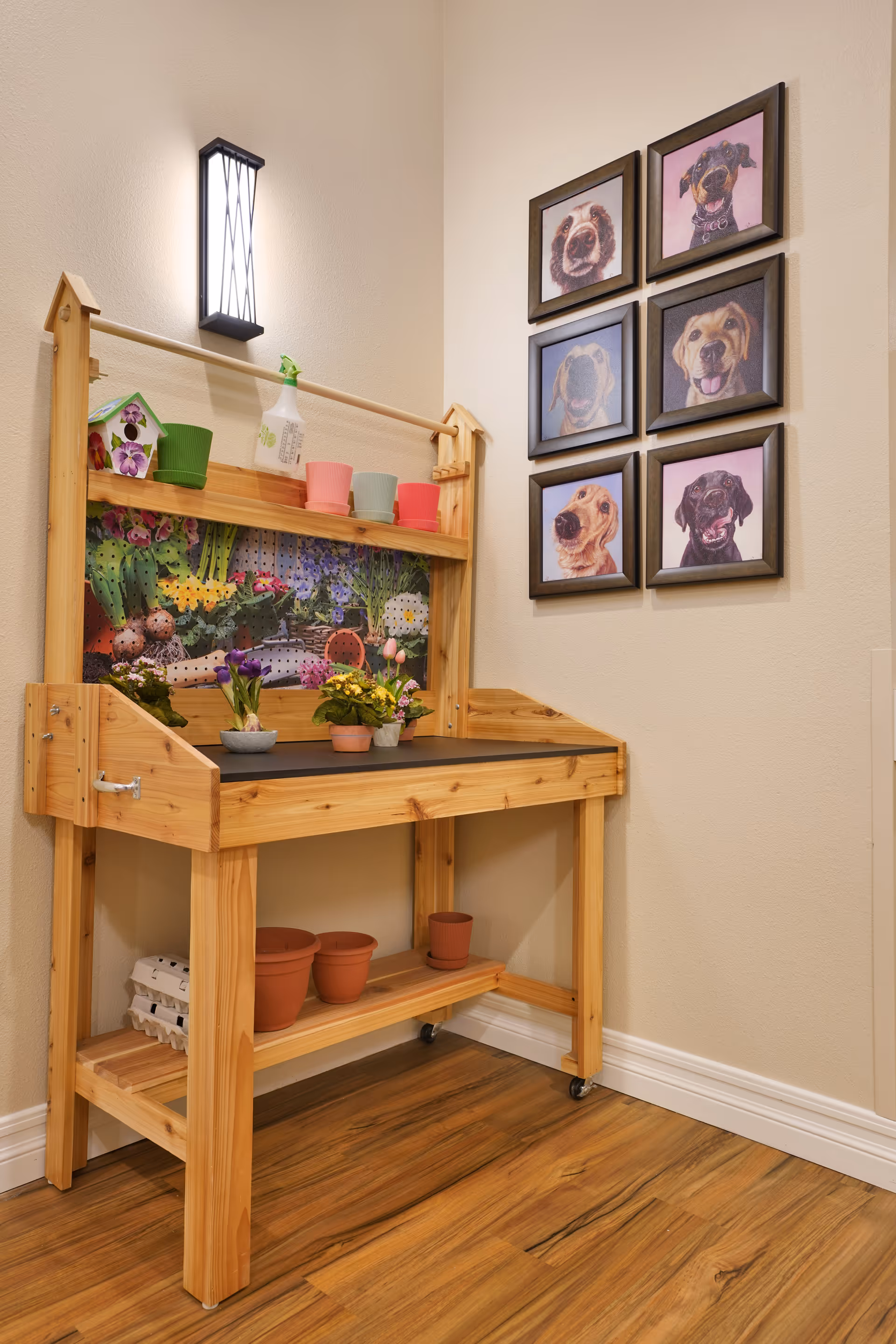 A wooden potting bench with small flower pots, a spray bottle, and a decorative birdhouse on top. The bench has a colorful floral backdrop and additional empty pots on the lower shelf. On the wall to the right, there are six framed pictures of dogs arranged in two columns of three. The room has light-colored walls and wood flooring.