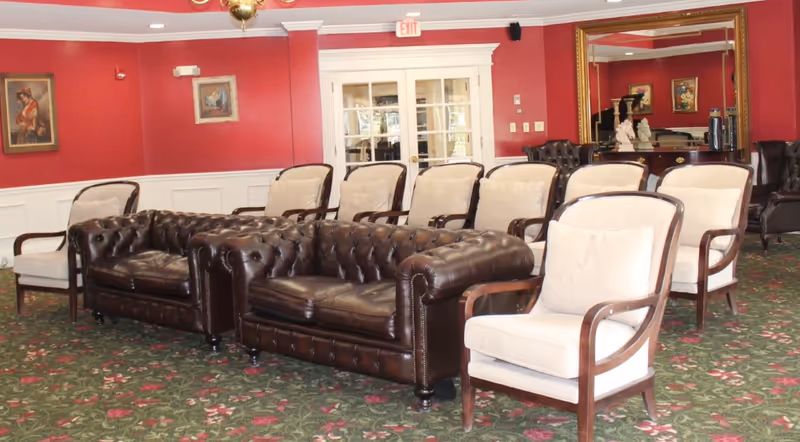 A sitting area with two dark brown leather tufted sofas and several beige cushioned armchairs arranged on a green floral carpet. The walls are painted red with white wainscoting, decorated with framed paintings. A large gold-framed mirror and a wooden sideboard are visible in the background, along with white double doors.