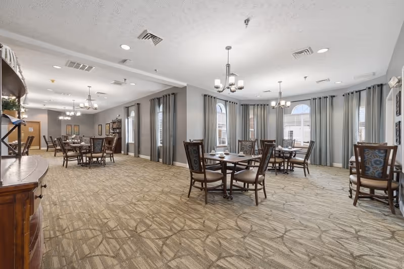 A spacious dining room with multiple wooden tables and chairs arranged neatly. The room features large windows with gray curtains, beige patterned carpet, and modern chandeliers hanging from a white ceiling. There are decorative shelves and framed pictures on the walls, creating a warm and inviting atmosphere.