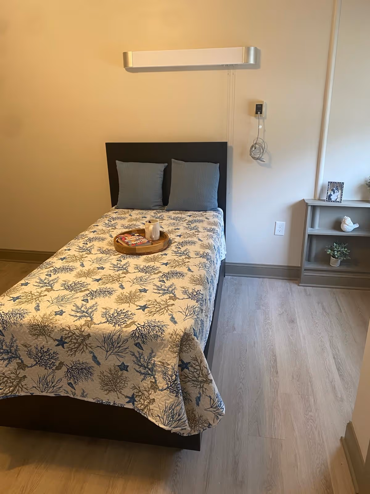 A single bed with a dark headboard and two gray pillows, covered with a white and blue patterned quilt featuring coral and starfish designs. On the bed is a wooden tray holding a white mug and a magazine. To the right of the bed is a small gray shelf with a framed photo, a white bird figurine, and a small potted plant. The room has light wood flooring and beige walls with a mounted light fixture above the bed and a call button on the wall.