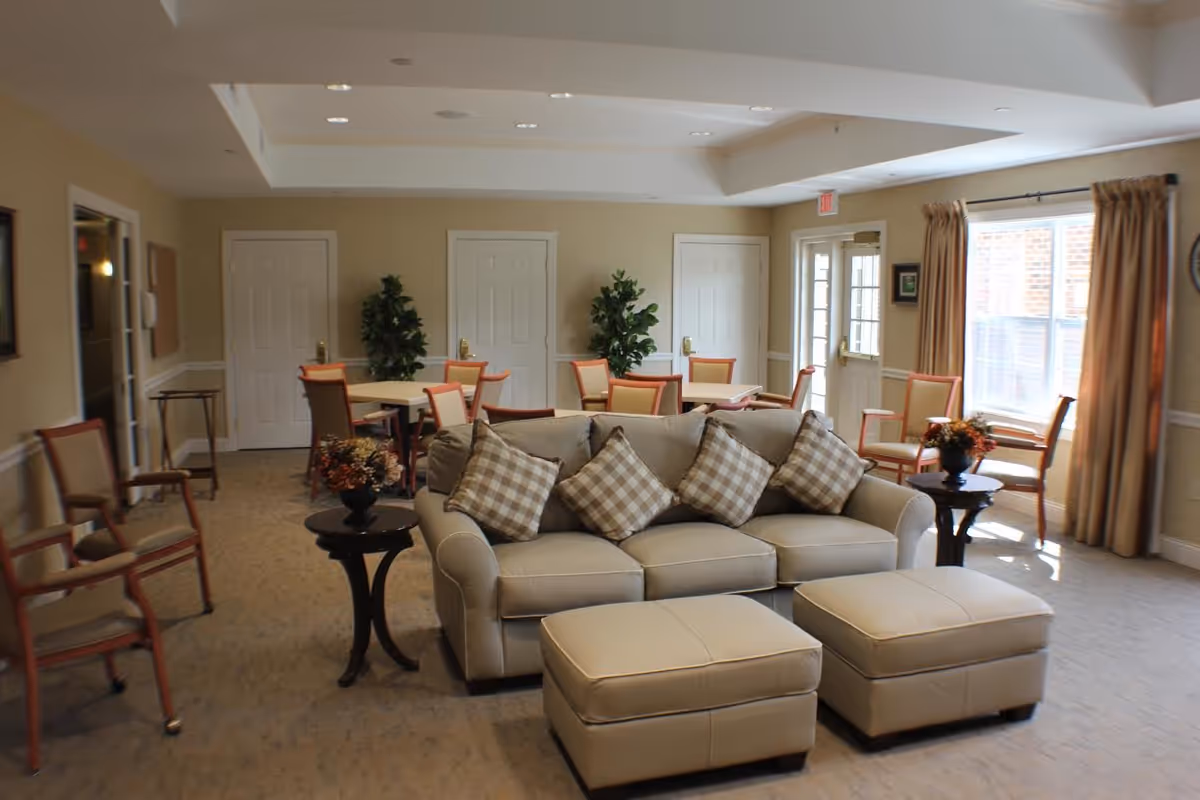 A bright communal living room with a beige sofa and ottomans, checkered throw pillows, side tables with floral arrangements, and dining tables and chairs near windows.