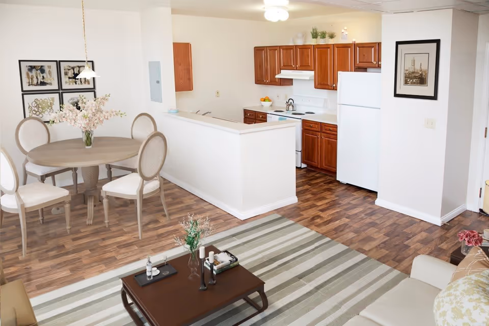 Interior view of a senior apartment showing a dining area with a round table and four chairs, a kitchen with wooden cabinets, white appliances, and a living area with a striped rug, coffee table, and beige sofa.