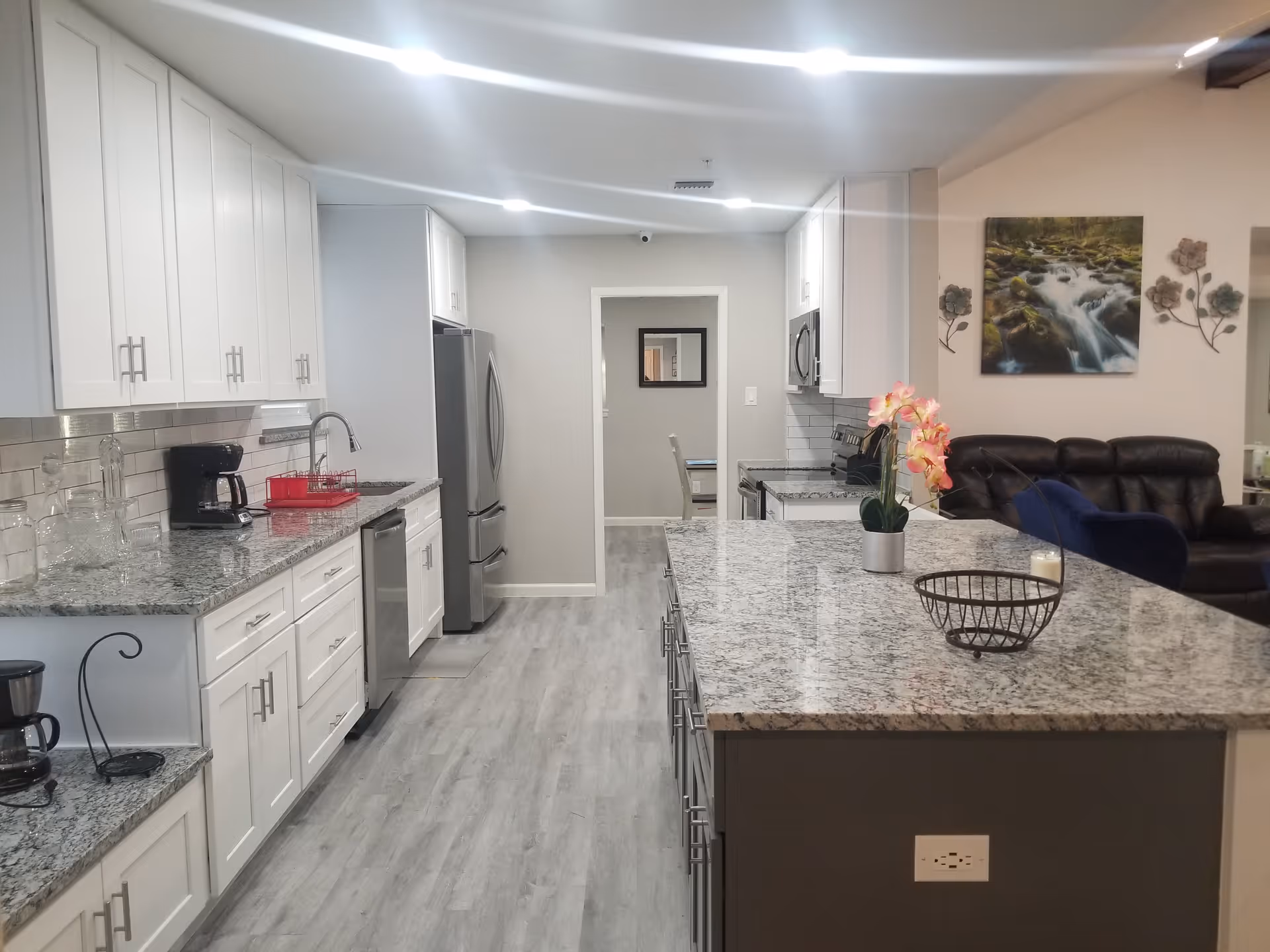 A modern kitchen with white cabinets, granite countertops, and stainless steel appliances including a refrigerator, dishwasher, and microwave. There is a large kitchen island with a flower pot and a wire basket on it. In the background, a living room area with a dark leather couch, a blue chair, and wall decorations including a painting of a flowing river and metal flower art is visible.