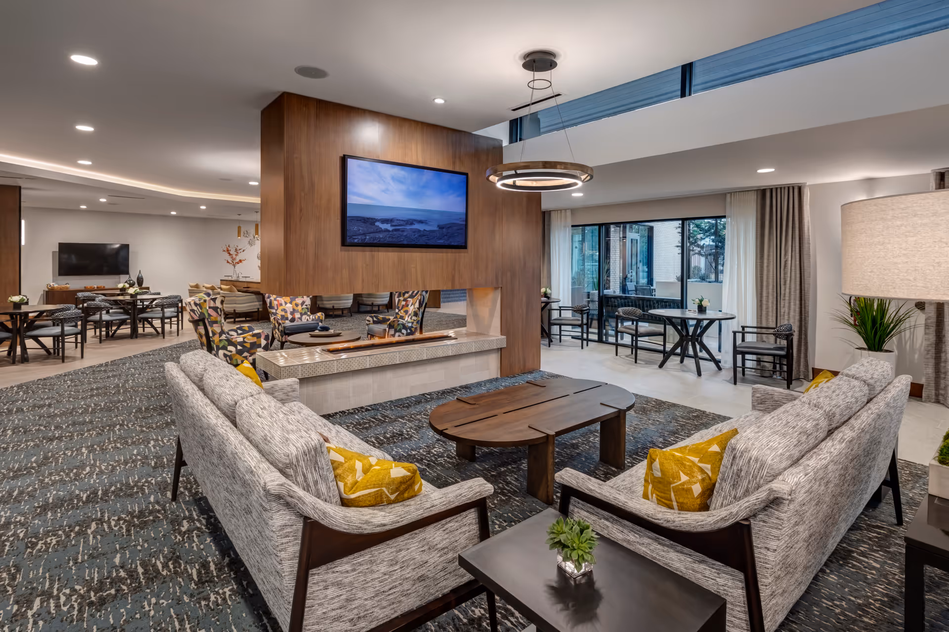 A modern and spacious living room area in a senior living facility featuring two gray sofas with yellow patterned pillows facing a wooden coffee table. A large wooden partition with a mounted flat-screen TV and a contemporary circular light fixture hangs from the ceiling. Behind the partition are colorful armchairs and tables, with additional seating and tables near large windows that let in natural light.