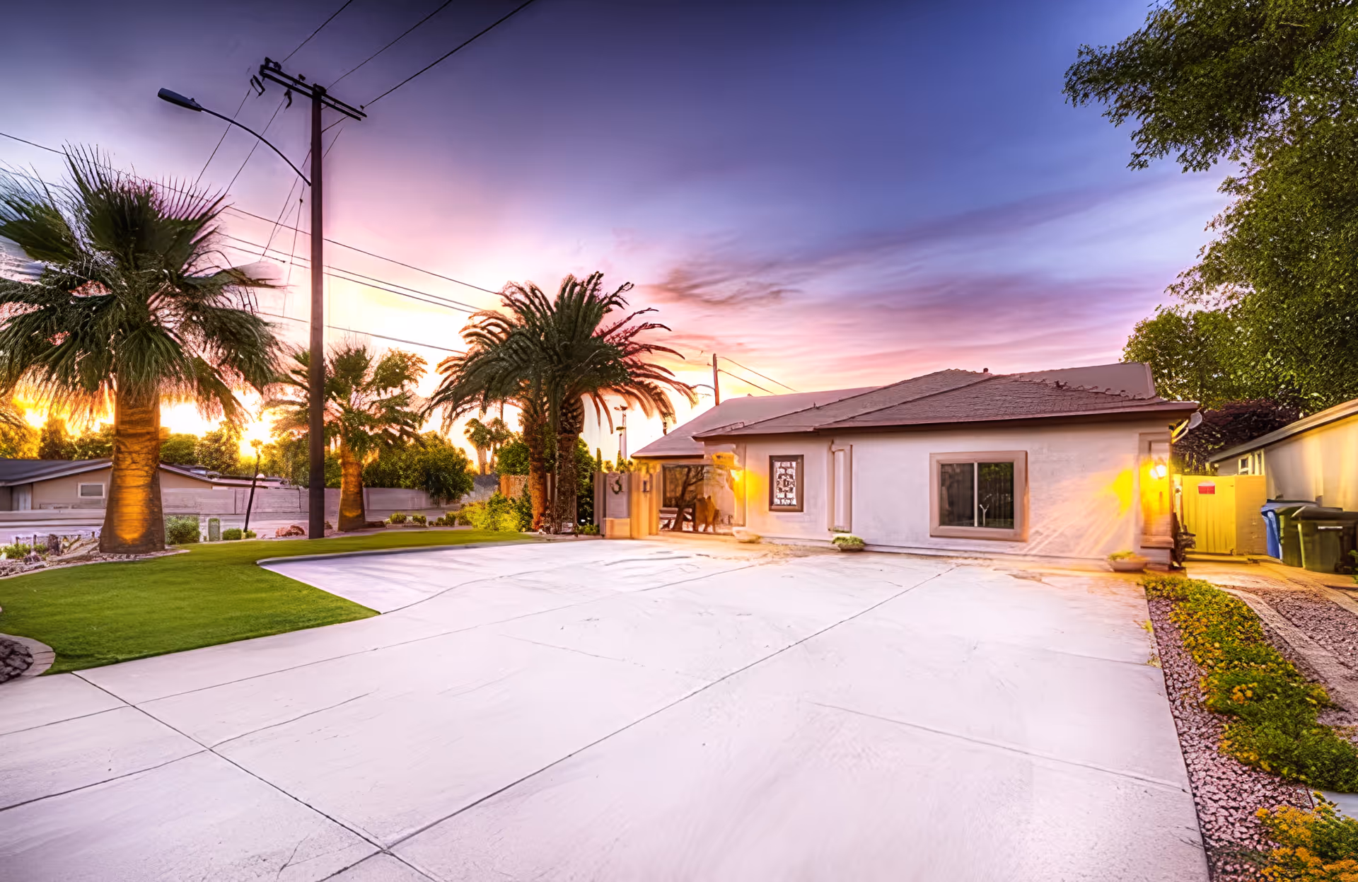 Exterior view of a single-story house with a large concrete driveway, palm trees, and well-maintained landscaping at sunset with a colorful sky.