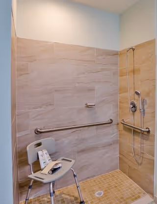 Accessible shower area with beige tiled walls and floor, equipped with a handheld showerhead, grab bars, and a white shower chair holding two bottles of shower products.