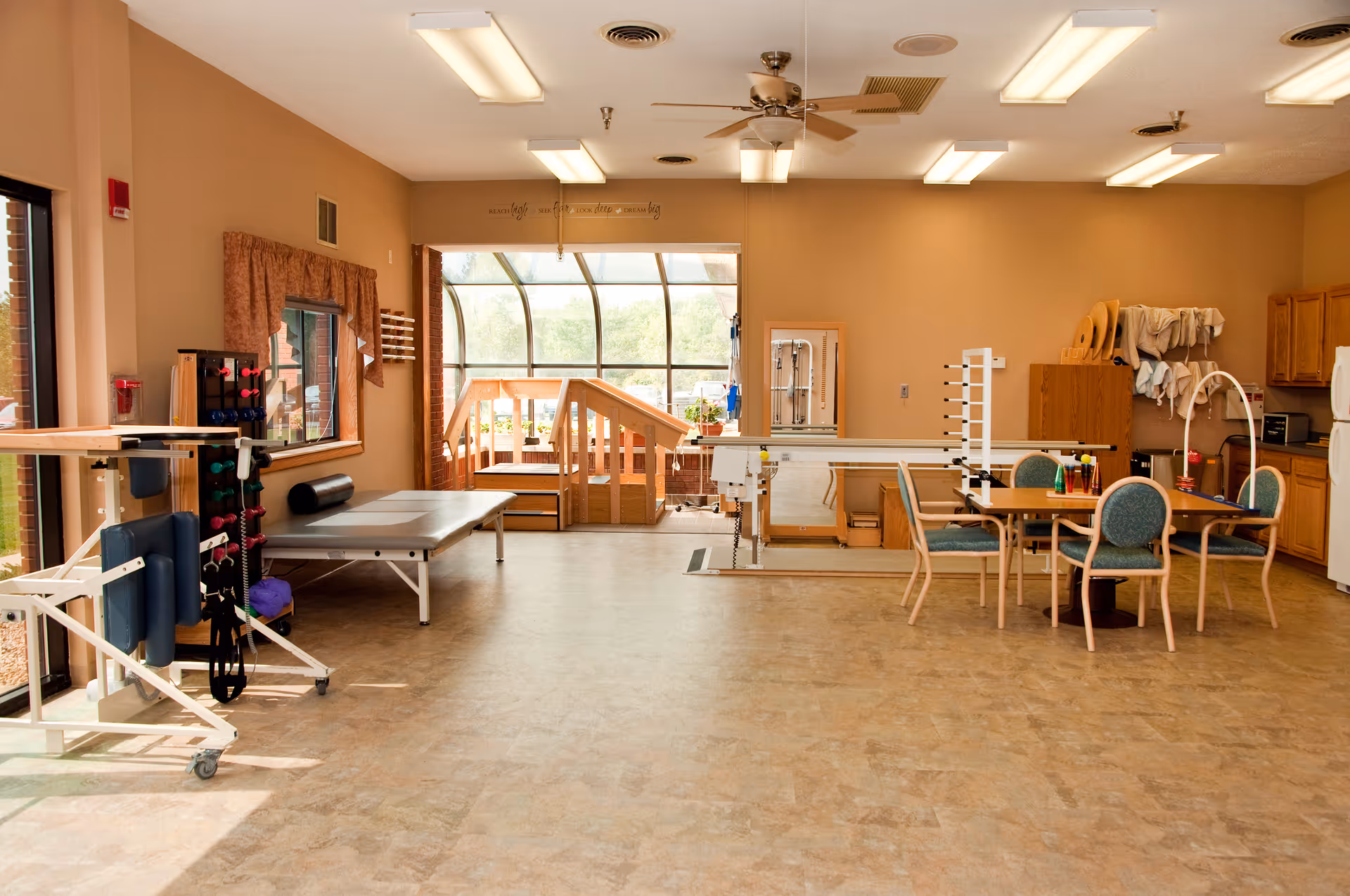 A spacious rehabilitation or therapy room with exercise equipment, a therapy table, chairs around a table, and a wooden ramp leading to a sunlit area with large windows. The room has beige walls, ceiling fans, and fluorescent lighting.
