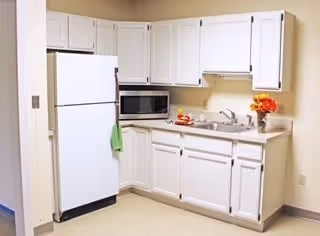 A small kitchen area with white cabinets, a white refrigerator, a microwave, a sink, and a countertop with a vase of orange flowers and a bowl of fruit.