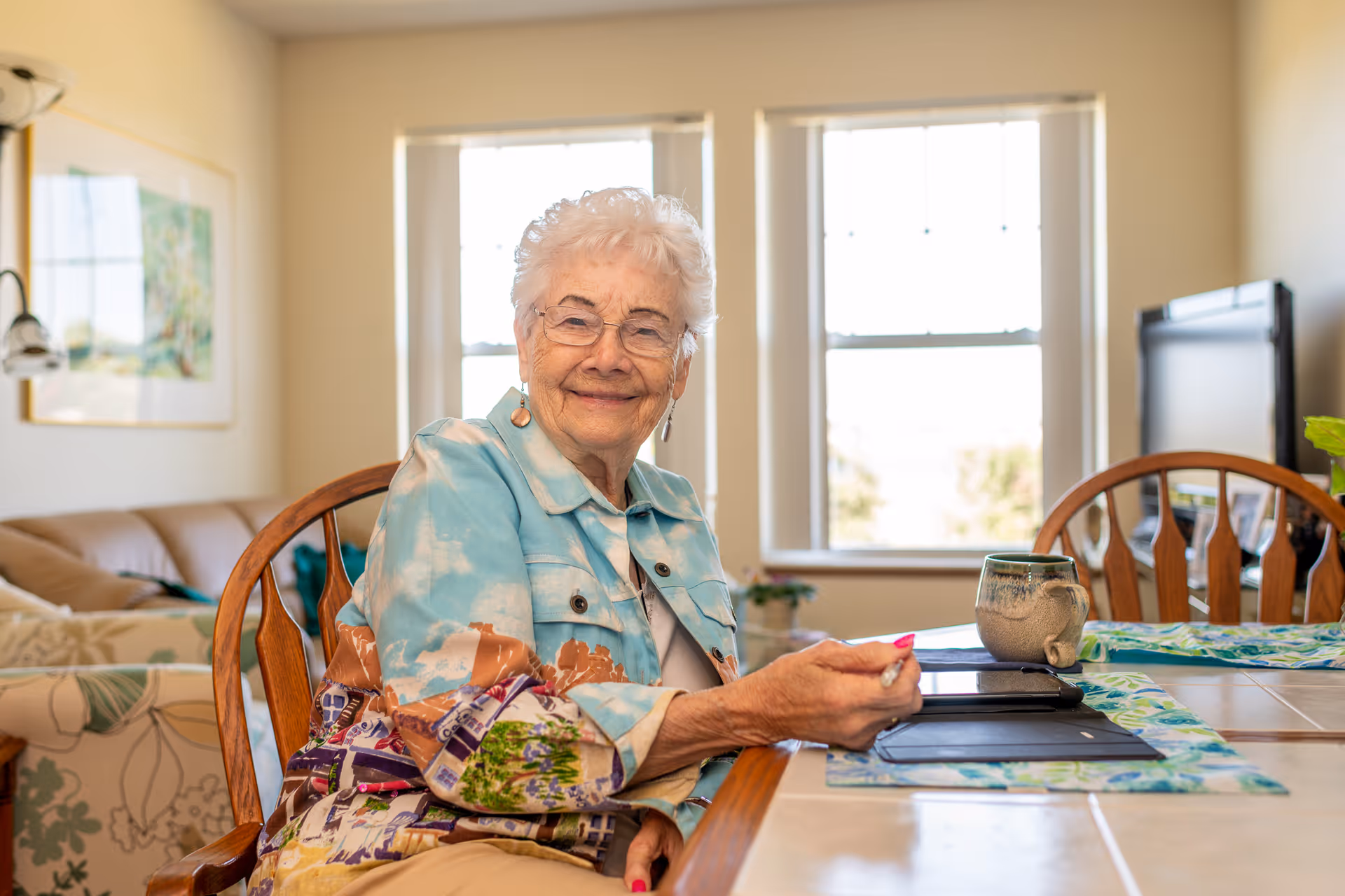 An elderly woman with white hair and glasses is sitting at a dining table in a well-lit room. She is smiling and wearing a colorful jacket. On the table in front of her are a mug, a tablet, and placemats. Behind her, there is a couch, a framed picture on the wall, and a window letting in natural light.