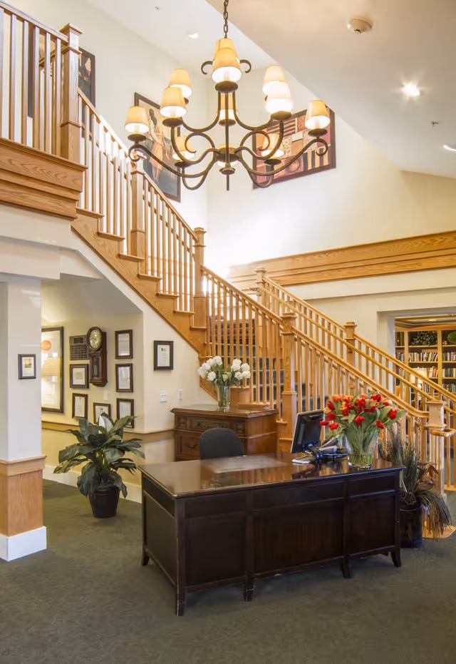 Reception area with a dark wooden desk, a computer monitor, and a vase of red flowers. Behind the desk is a wooden staircase with railings leading to an upper floor. The walls are decorated with framed pictures and a large chandelier hangs from the ceiling. There are also plants and a small wooden cabinet with white flowers on top.