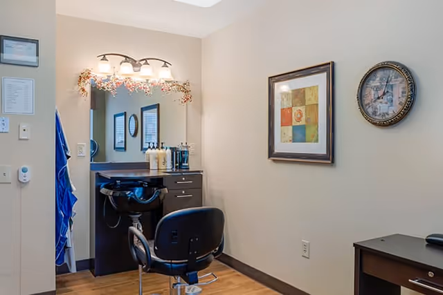 Interior view of a salon area in a senior living facility with a black salon chair in front of a black sink and countertop. Above the sink is a large mirror with a light fixture and decorative garland. On the wall to the right, there is a framed colorful artwork and a round clock. The room has beige walls and wood flooring.