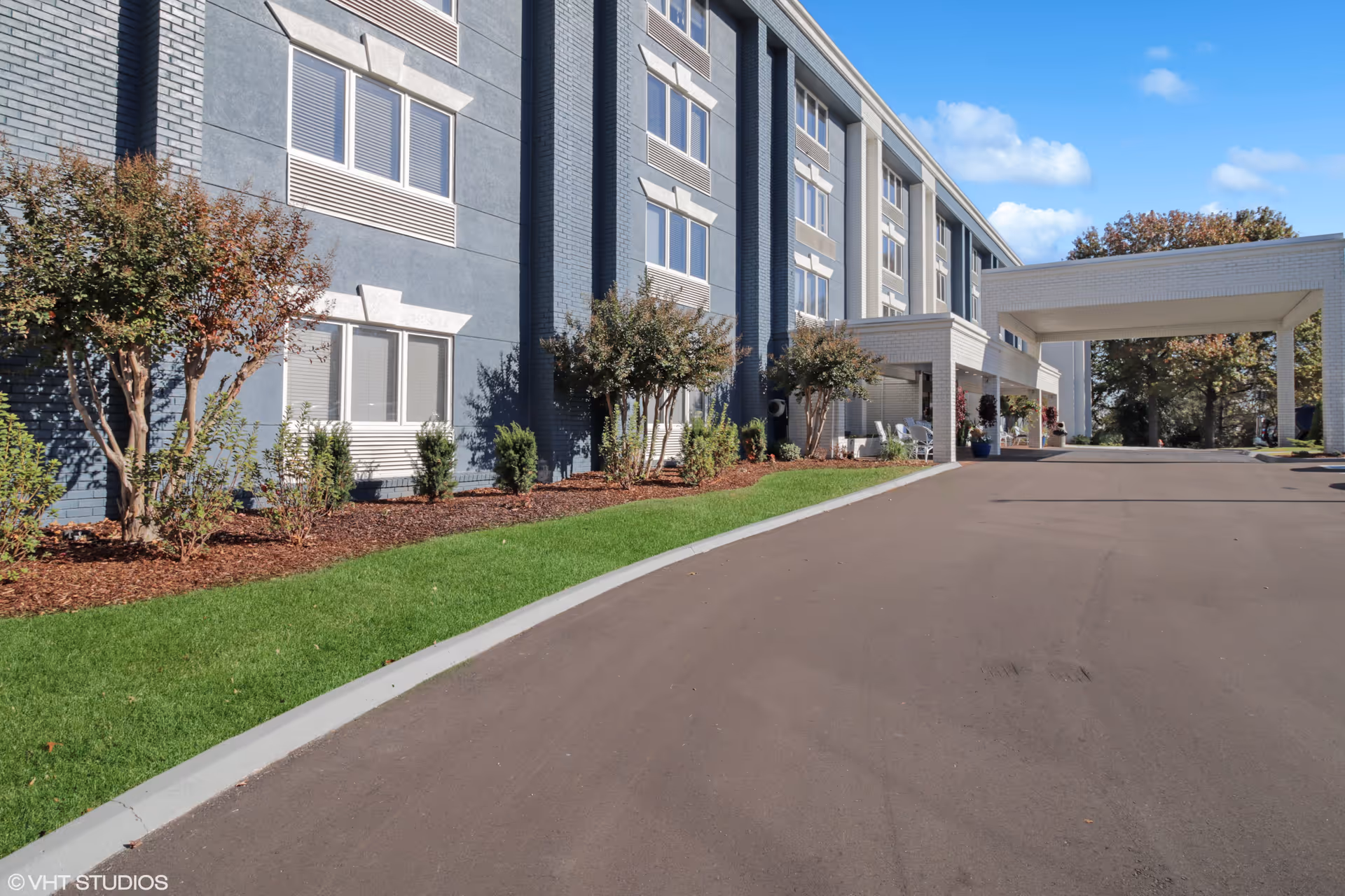 Exterior view of a multi-story senior living facility building with a covered entrance driveway, landscaped bushes and trees along the side, and a clear blue sky.