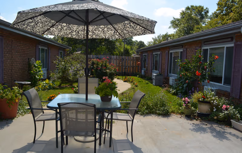 Outdoor patio area with a glass table and four chairs under a large patterned umbrella. The patio is surrounded by two single-story brick buildings with windows and flower beds filled with various plants and flowers. A concrete pathway leads through a grassy garden area between the buildings under a partly cloudy sky.
