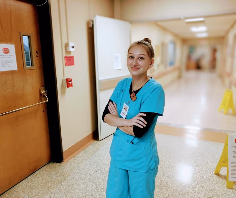 A young female healthcare worker in turquoise scrubs stands in a hospital hallway with her arms crossed, smiling at the camera. The hallway has beige walls, a wooden door with a stop sign, and a wet floor caution sign in the background.