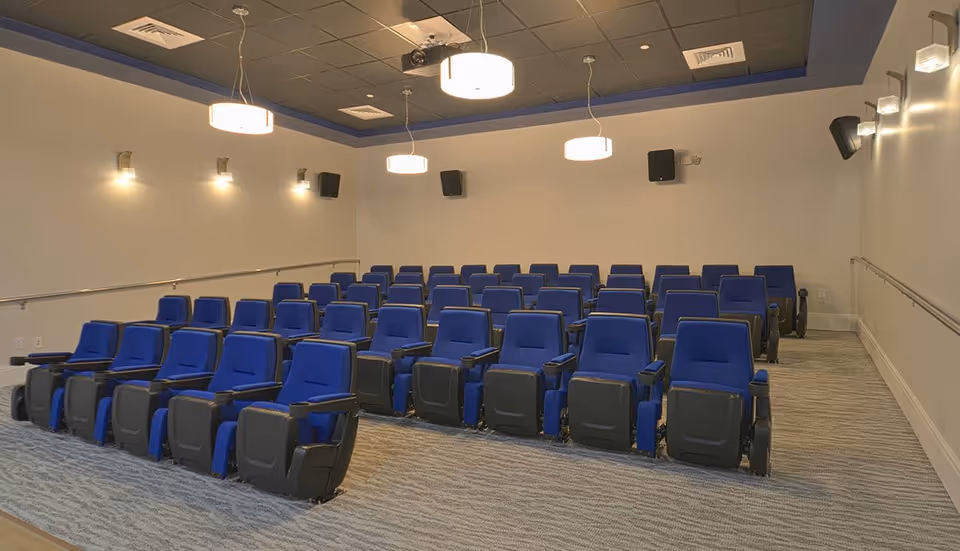 Small screening room with rows of blue theater seats, overhead pendant lights, wall speakers, and a ceiling-mounted projector.