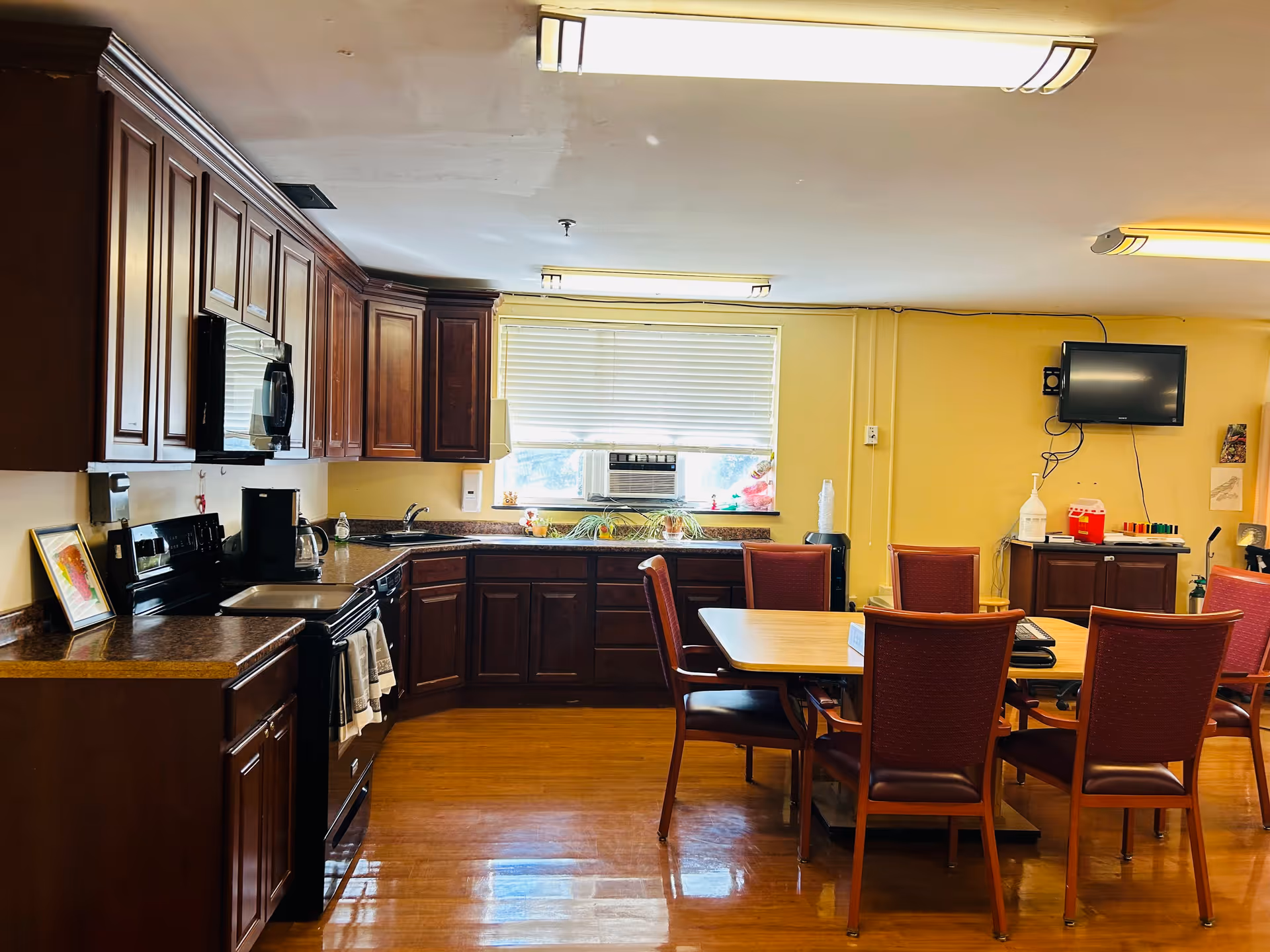 Interior view of a kitchen and dining area in a senior living facility. The kitchen features dark wooden cabinets, a stove, microwave, coffee maker, and a sink under a window with blinds and an air conditioning unit. The dining area has a wooden table surrounded by six red cushioned chairs. A wall-mounted TV and various items are visible on a cabinet against a yellow wall.